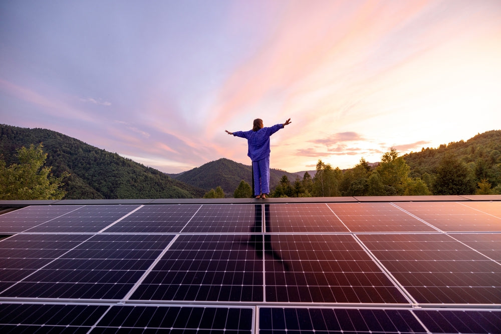 Person standing on a solar panel with a scenic mountain landscape and sunset in the background