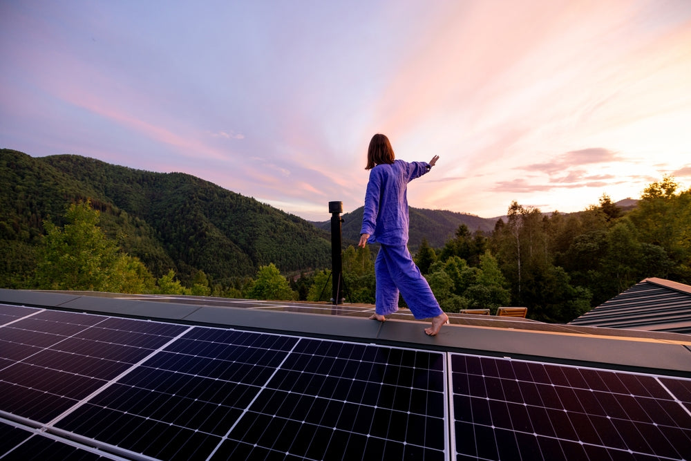 Woman walking barefoot on rooftop solar panels during sunset with scenic mountain and forest view.